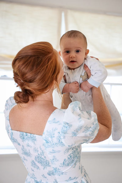 Therese's is wearing the Lan Midi Dress with nursing zippers in Blue La Mere Toile, showing off the smocked back, as she lifts up her baby boy 