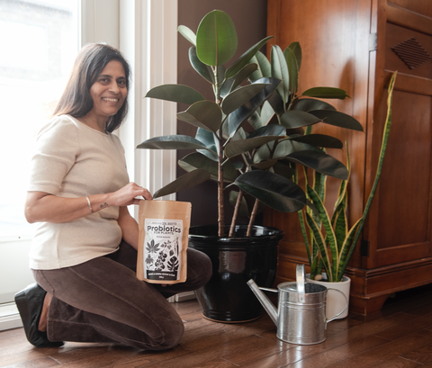 smiling woman kneeling beside houseplants in front of window in living room holding a small bag of probiotics for plants worm manure