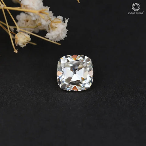 [Close-up of a cushion-cut moissanite gemstone on a dark background, with delicate white flowers in the corner.]-[ouros jewels]