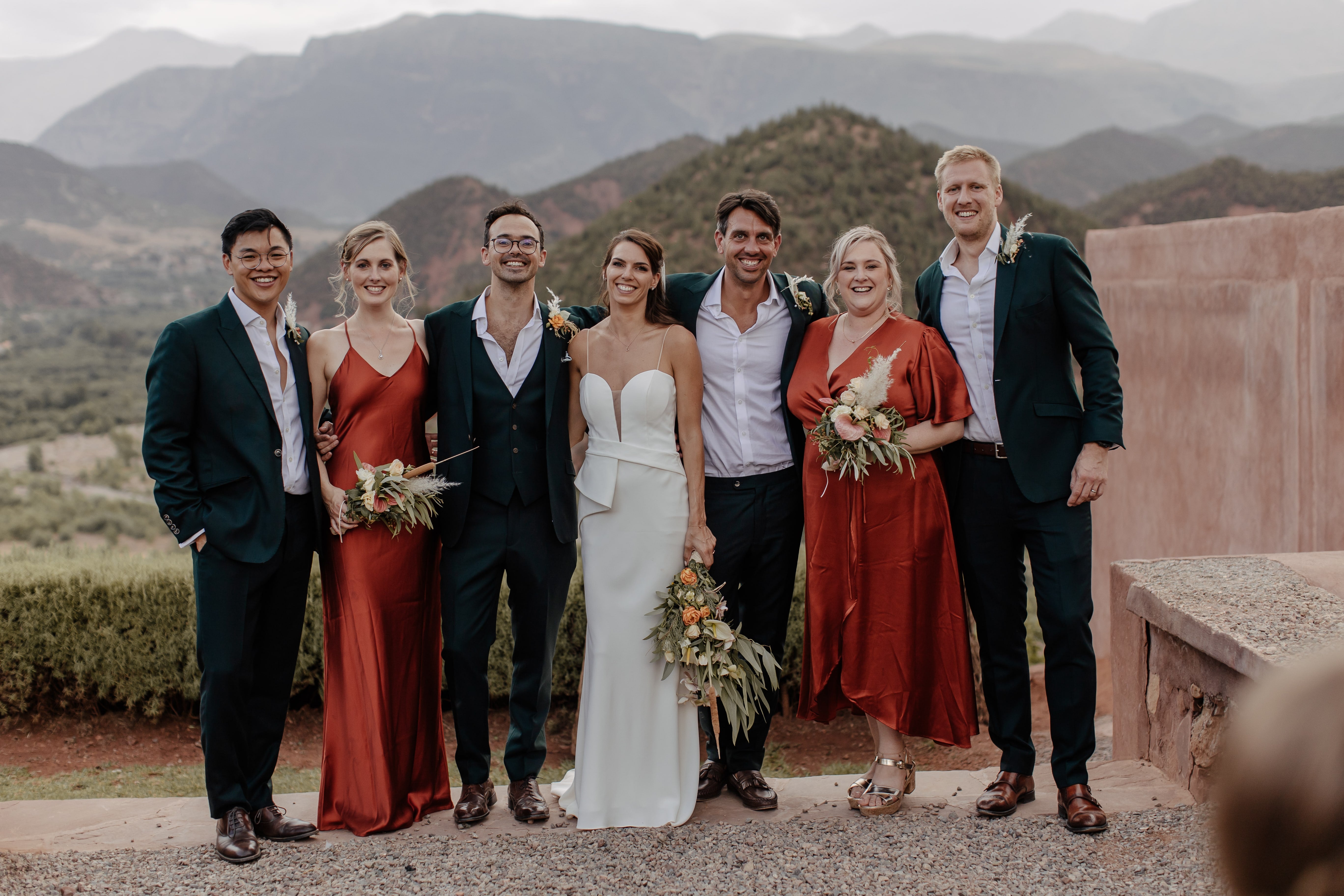 Bridal party of 7 people, including bride and groom and 2 bridesmaids and 3 best man, standing in a line in outside location with a skyline of hills in the background.