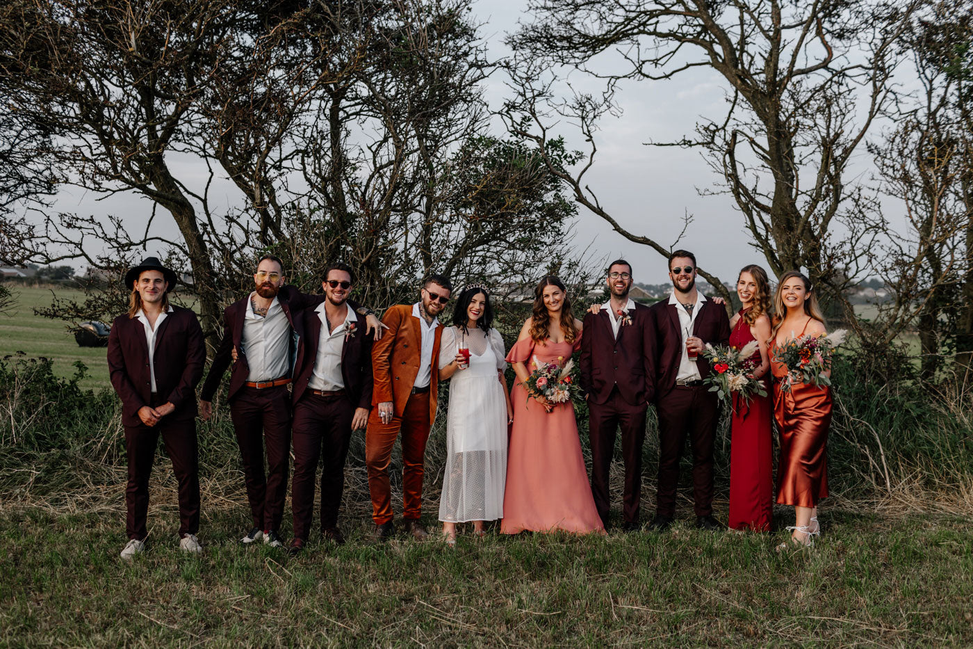 Bridal party with bride and groom, bridesmaids and best men. Bridesmaids in a mix of burnt orange satin, dark red and coral dresses.