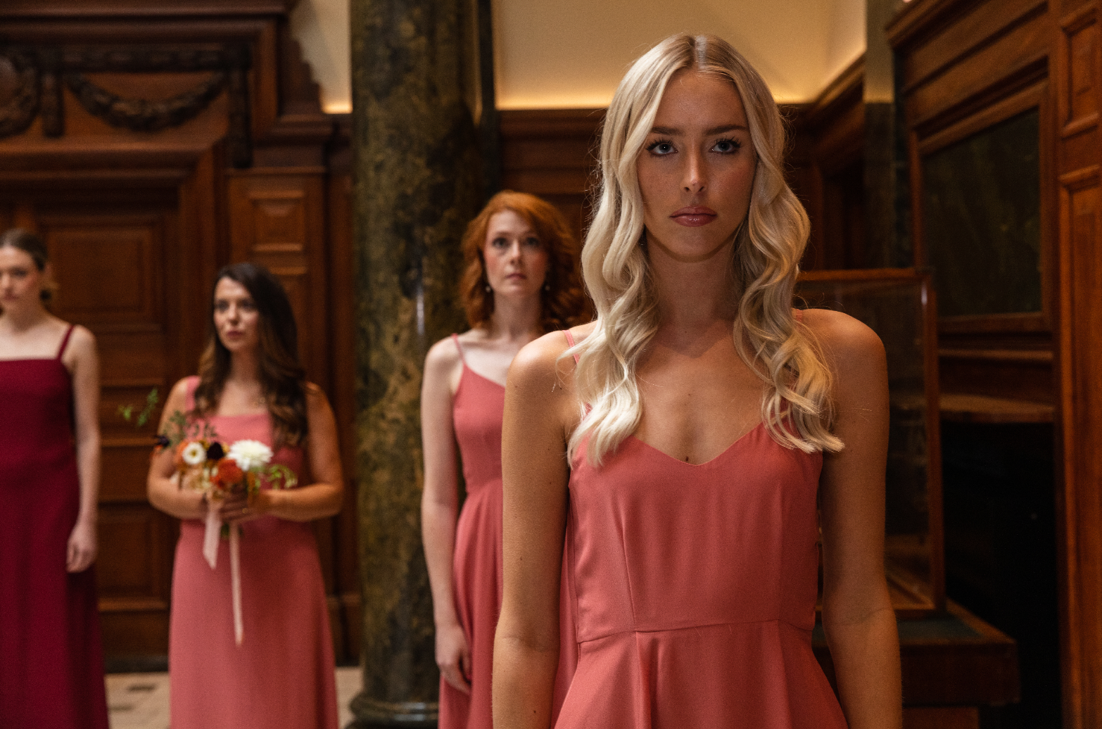 A group of girls standing in the foyer of the Town Hall Hotel in a regimented formation, all in a mix of red, coral and orange bridesmaids dresses, some holding bouquets of flowers.