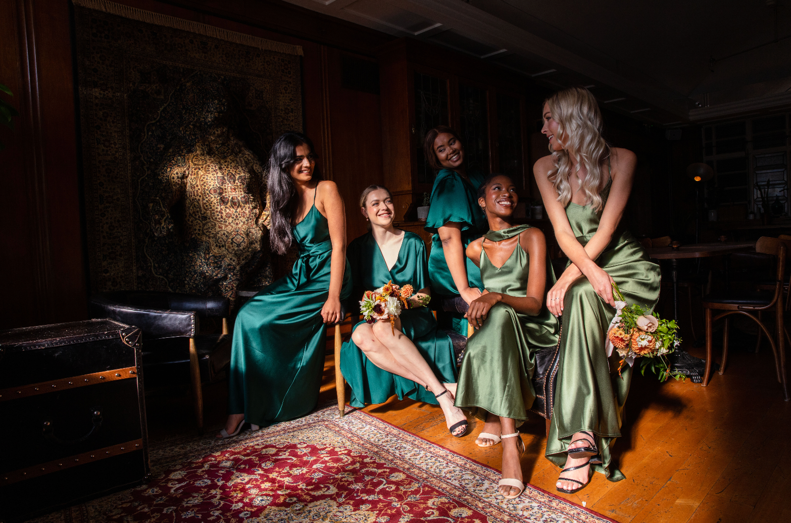 Five girls in a dark room with the sun shining through the window on them, all sitting down at different levels on a chair and sofa in different shades of green satin bridesmaids dresses. One style is a wrap midi length and the other styles are strappy slip with a v neck and waist sash.