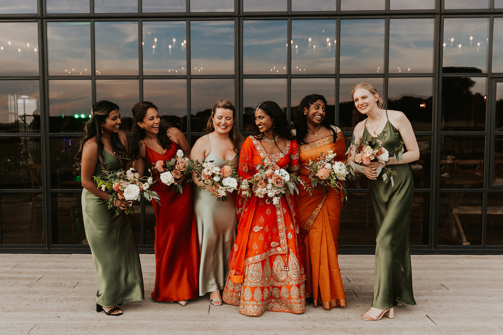A bridal party of 6 females including the bride, dressed in orange and green bridesmaids dresses with the bride in an orange South Asian Lehenga.