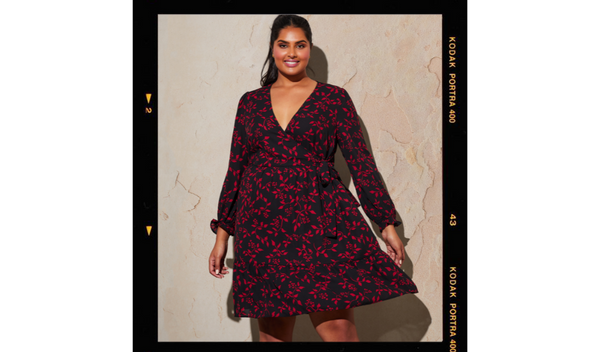 Pictured: model Shanaya smiling wearing the Shadow Floral Dress. The dress is black with red leaves and flowers, with long sleeves and a v neckline 