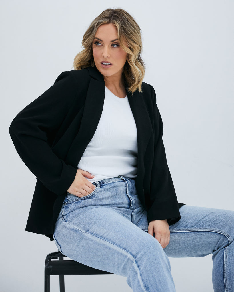 Pictured: Female model looking out of shot, Jess has shoulder length blonde hair and she is sitting on a stool. One hand is in her jean pocket. She is wearing light wash jeans, a white top and The Megan Blazer - a black boxy female plus size blazer.