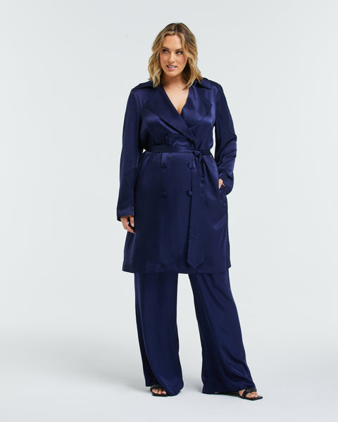 Pictured: Curve model poses for the camera. She is wearing a navy blue satin trench coat and a matching pant