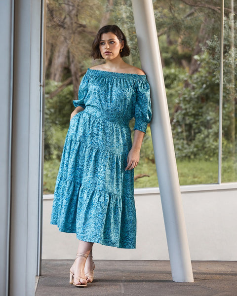 Pictured: Model leaning on a pole wearing the Sorrento Top and Sorrento Tiered Skirt by Estelle Clothing. Both pieces are light blue with a dark blue floral outline print.