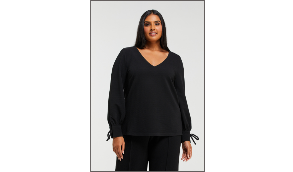 Pictured: Female curve model is wearing the Clementine Top. The top is a women's plus size black long-sleeved top with loose fitting sleeves with cuff tie detailing.