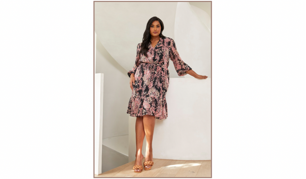Pictured: A female curve model is wearing the Harlequin Blooms Dress by Estelle. The dress is a women's plus size long sleeve floral dress.
