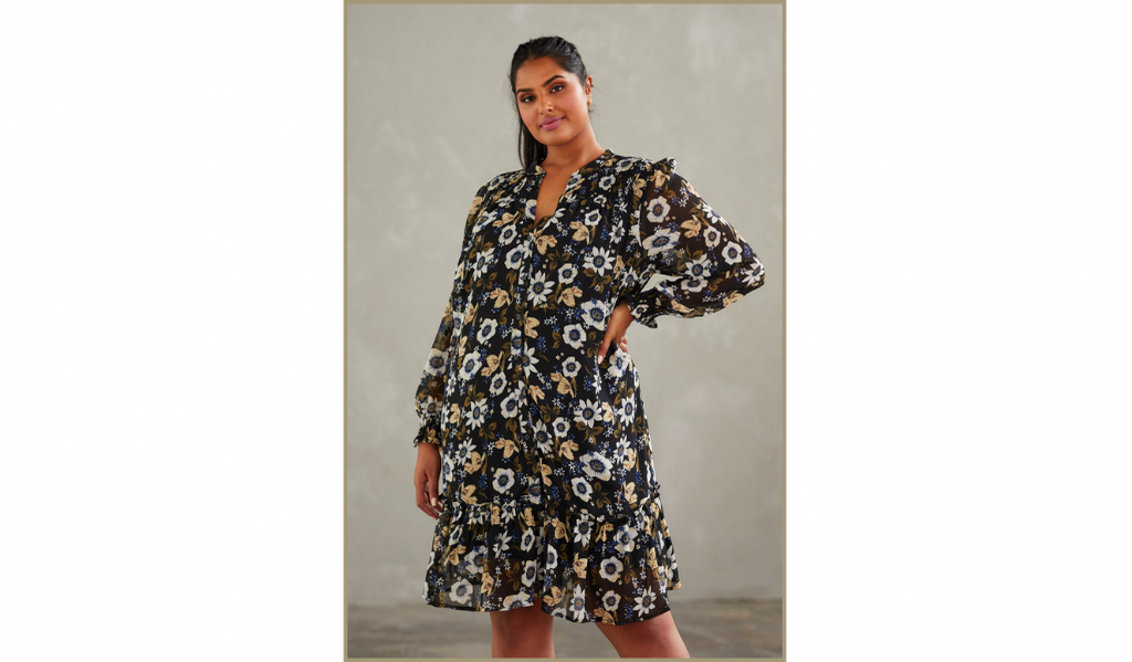 Pictured: A female curve model wearing the Delfina Dress by Estelle. The Dress is a loose fitted long sleeved floral printed dress that ends just above the knee. 