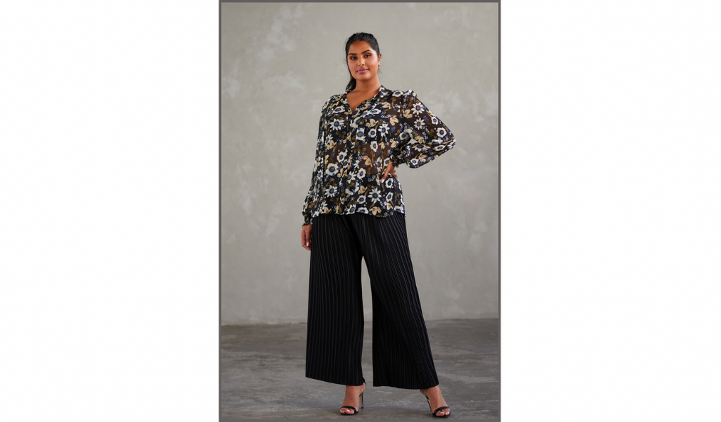 Pictured: A female curve model is standing posing for the camera. She is wearing the Vespa Pant by Estelle in Black. The Pant is a women's plus size loose fitting wide leg pleated pant