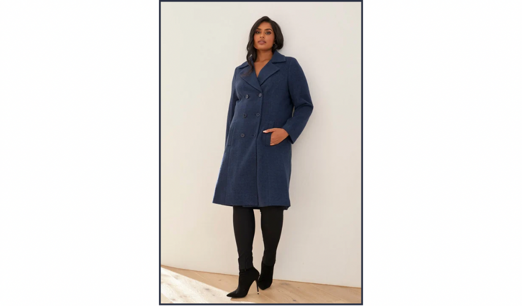 Pictured: A female model is leaning against a wall. She is wearing black boots, black tight fitting pants and the Fleet Coat by Estelle. The coat is a women's plus size navy knee length coat.