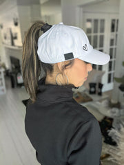 Image shows a female-presenting model wearing a white baseball cap with a high ponytail