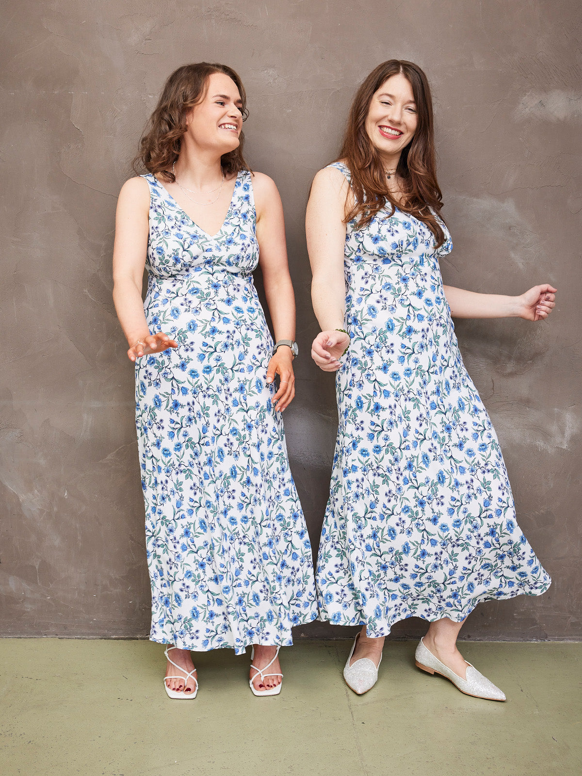 Two smiling women in matching floral dresses leaning against a textured wall.