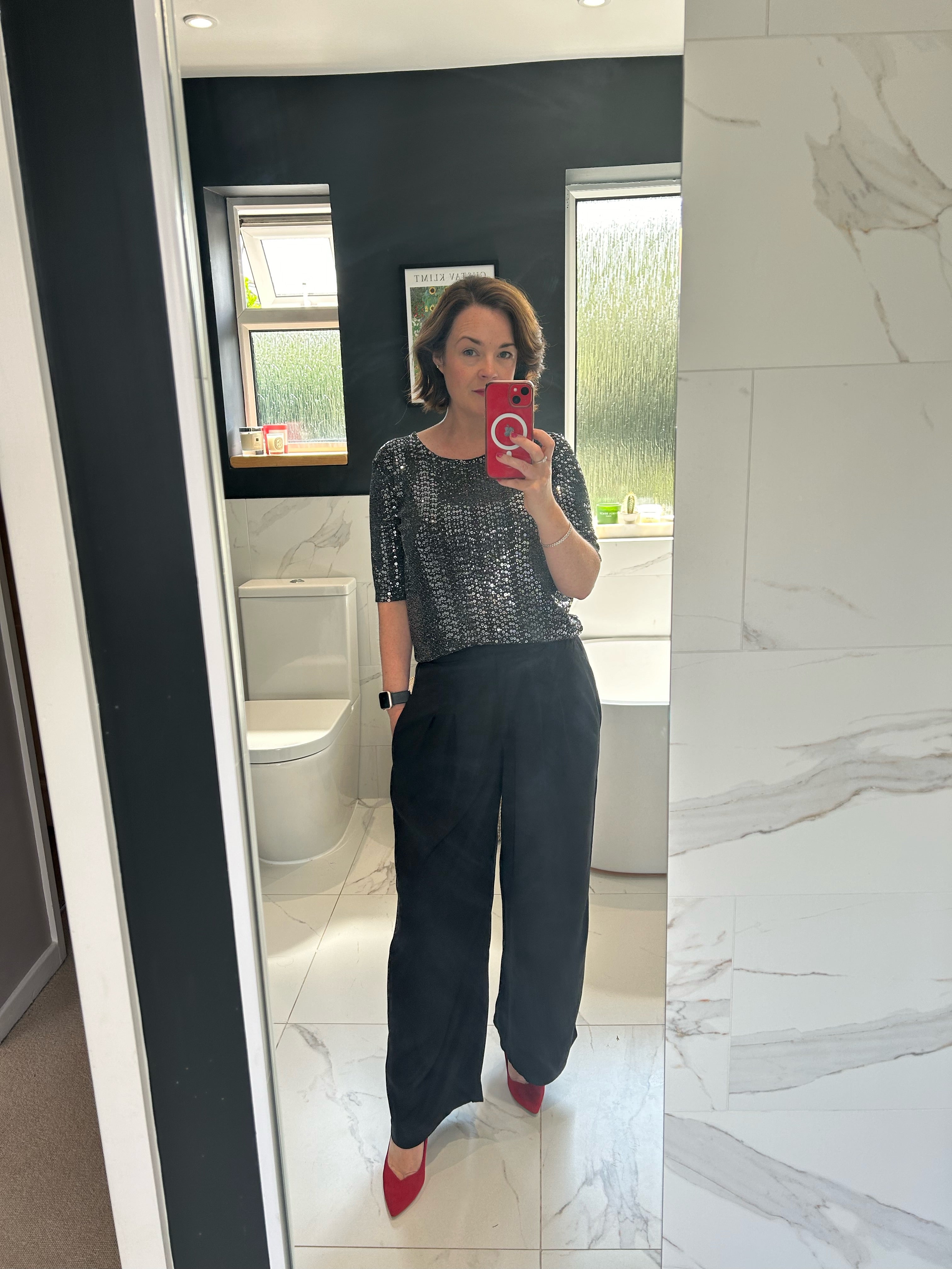 Woman taking a mirror selfie in a bathroom, wearing black pants, a sparkly top, and red shoes.