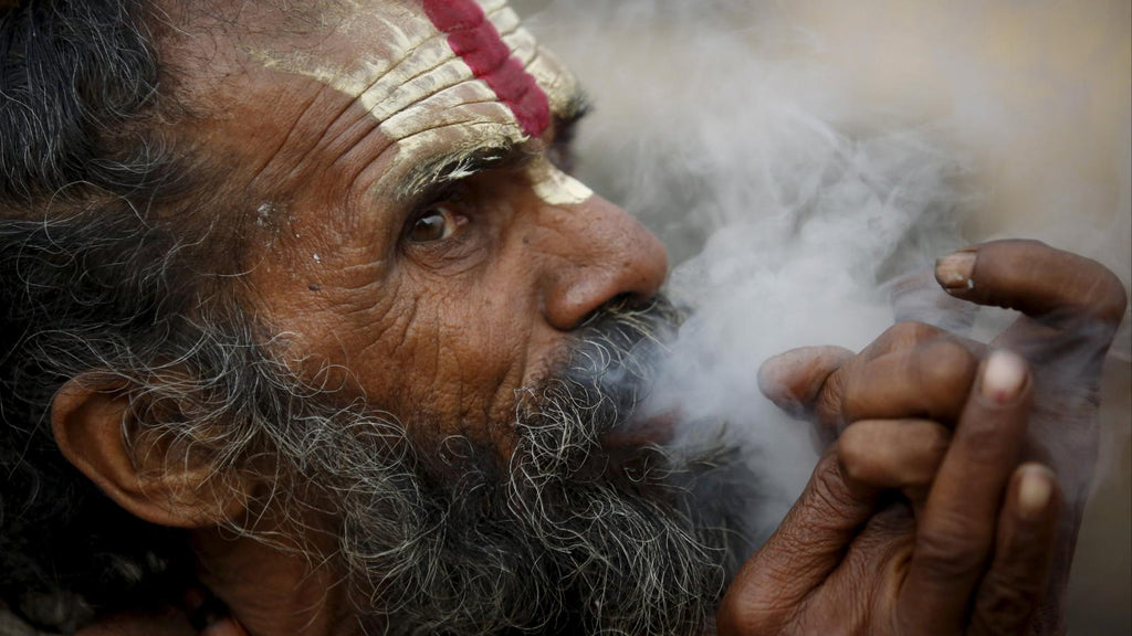Older Indian man with red make up paint on his face smoking a cannabis pipe.