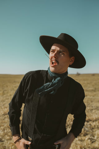 Cowboy in black waist coat and hat with blue background in a field with straw in his mouth.