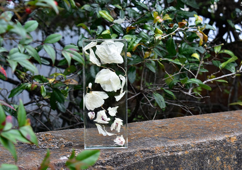 A 9'' x 4'' transparent brick made of resin sits on a cement slab. Inside the transparent brick dried orchids are arrange in a way that makes them appear to be floating away.