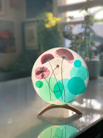 Poppies and green and blue disks are suspended in a 7'' diameter circle of resin. The artwork is position on a wooden stand, illuminated from behind by the afternoon light.