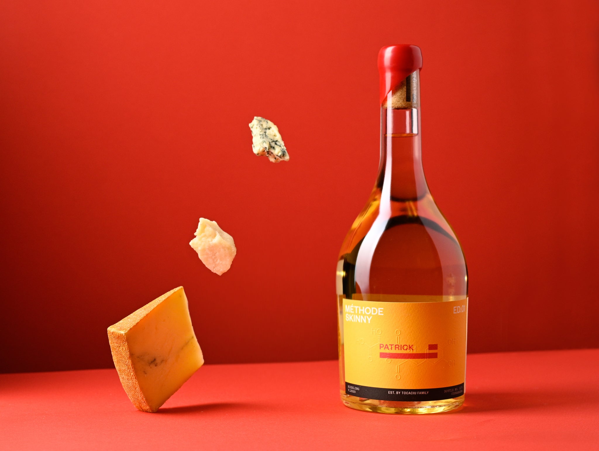 A bottle of white wine with a yellow label against a bright red background, next to 3 falling chunks of cheese.