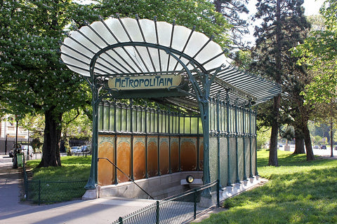 Paris metro station entrance at Porte Dauphine designed by Hector Guimard for the 1900 Exposition universelle. It is a large, glass structure atop a subway with stairs leading underground. The glass is formed in the shape of a fan, with black lead holding the panes together. The sign says 'Metropolitan' in a dark blue or green. There are dark orange decorated panels on the walls.