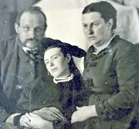 Example of death photography; a group of three people, two elder, a man and a woman with soft, slightly blurred features. They are positioned either side the body of a younger, dark haired woman with open eyes. She is the main feature of the portrait and her features are in focus.