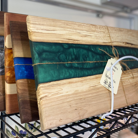 Three wood and epoxy cheese boards stacked on a shelf, for sale, in various colours such as green, blue, and gold.