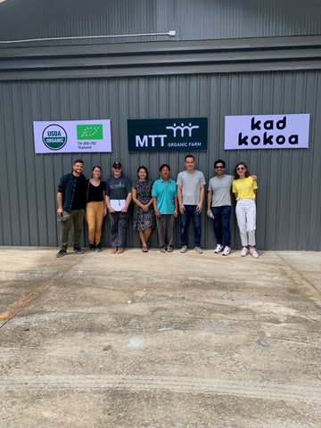 Image depicting a diverse team of farmers from MTT Farm and representatives from Kad Kokoa gathered together at the Kad Kokoa and MTT Farm factory in Chiangmai. The group, showing a mix of men and women of various descents, is smiling and standing proudly in front of the factory, which displays both the Kad Kokoa and MTT Farm logos. The factory setting includes cacao processing equipment in the background, illustrating the collaborative work environment where cacao beans are transformed into high-quality chocolate products.
