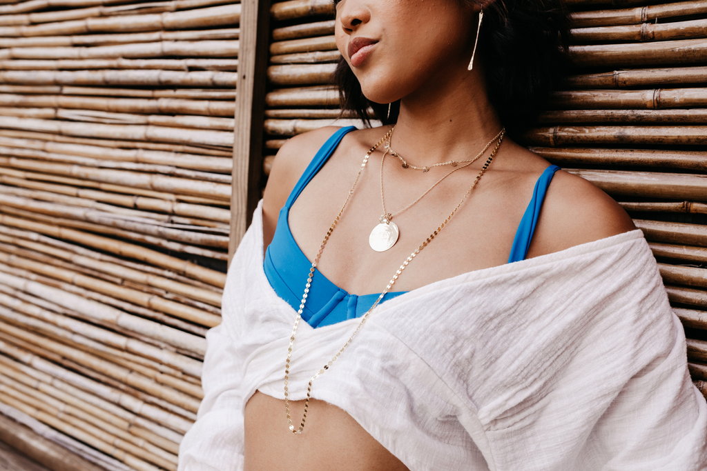 Model leans back in a blue bikini with white beach coverall wearing Minimalist Stick Earrings, 14k Gold Filled Disc Necklace, Crystal Medallion Necklace and Mykonos Layer Chain.