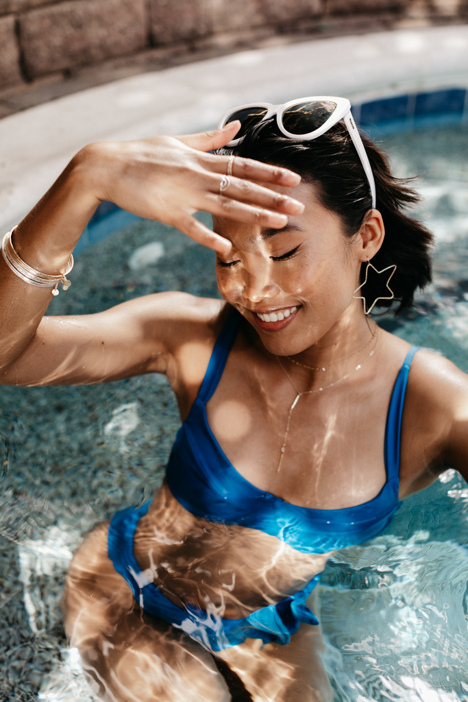 Model in a blue bikini uses her hand to shade the sun from her eyes wearing a Layering Chain, Bar Lariat Necklace, Bangle Bracelet Set, Minimalist Stacking Band Rings, 14k Gold Filled Arch Ring and Star Hoop Earrings.