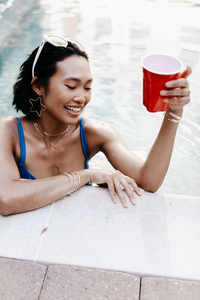 Model celebrates the 4 July in the pool holding a red cup and wearing a blue bikini, white sunglasses, layering Chain, Minimalist Bar Lariat Necklace, Bangle Bracelets and Water Resistant Star Hoop Earrings.