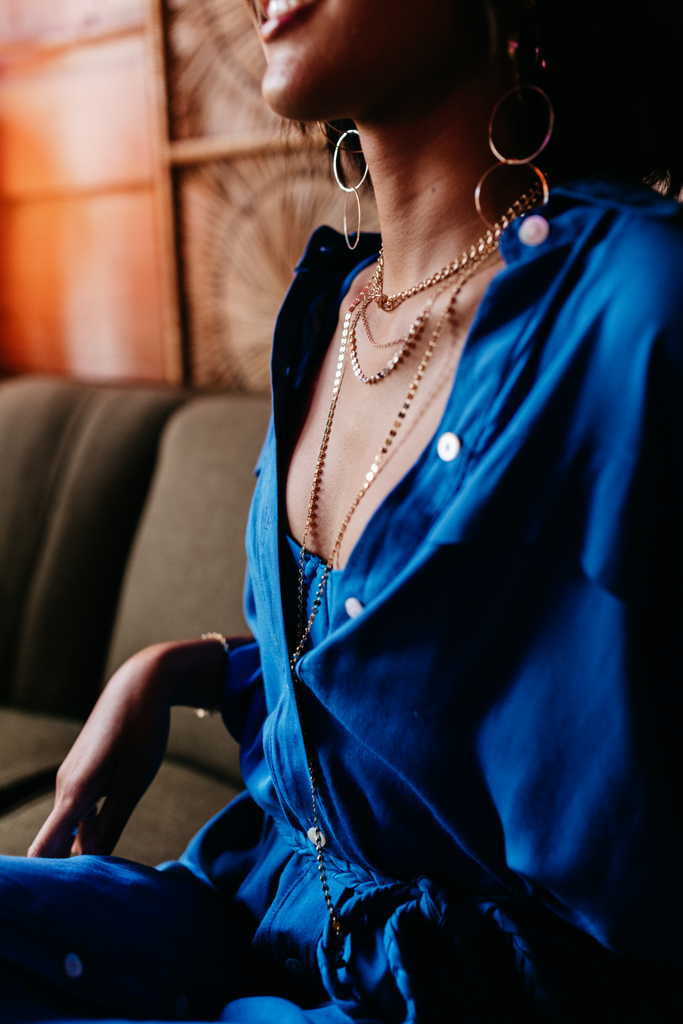 Model laughs in the bar wearing a blue button up shirt over a blue bikini with Lightweight Gold Sunnies Hoop Earrings, Chain Necklace with Toggle Clasp, Mykonos Layering Necklace, and Mykonos Long Layer Chain.