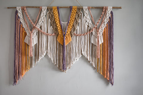 A large macrame wall hanging with day break colors - purple, pink, orange, yellow, tan and white. The layers and spirals sweep and overlap from a bamboo dowel. It is majestic.