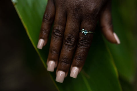 a dark skinned hand hovering above greenery highlights a pear and marquise cut moi et toi style hammered ring in silver