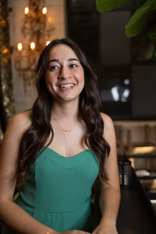 a woman in a green dress with long dark wavy hair stands in front of a chandelier wearing a gold and diamond necklace