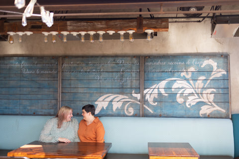 two women sit looking at each other on a restaurant patio with French poetry written on the wall behind them.