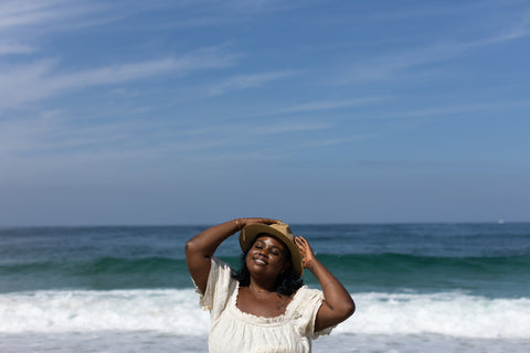 A black woman wearing a sun hat and gold jewelry smiles and closes her eyes, looking towards the sun with the ocean behind her.