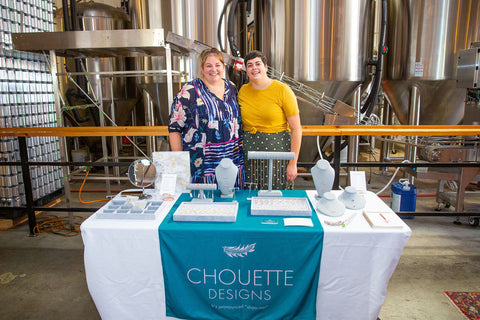 Chouette Designs founders Ashley and Marine stand behind their booth at a pop up event with all the jewelry on display.