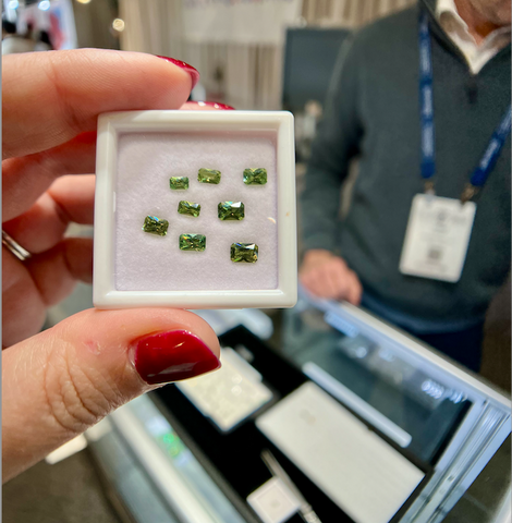 A hand holds up a package of sparkling green radiant cut demantoids with a trade show booth out of focus in the background