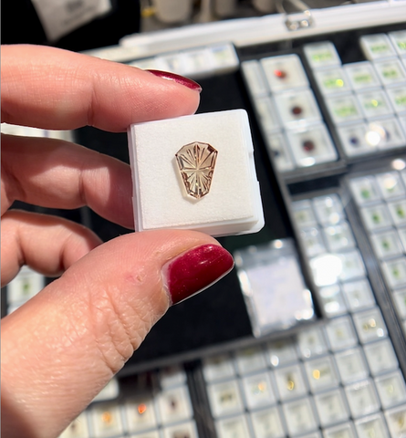 a hand holds up a shield fantasy cut Oregon Sunstone with out of focus gemstone displays in the background