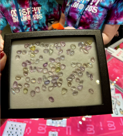 A large gemstone display with jelly-bean-esque pastel hued sapphires. Two booth employees are in the background and "Misfit Diamonds" can be clearly read on their tie-dyed t-shirts.