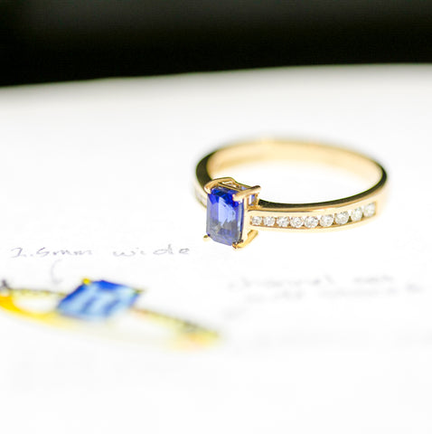 An emerald cut sapphire ring with channel set diamonds in yellow gold sits on a sketch of the ring.
