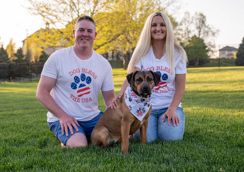 Foster dog mom and dad sitting in grass with brown foster dog wearing 4th of July theme shirts and bandana
