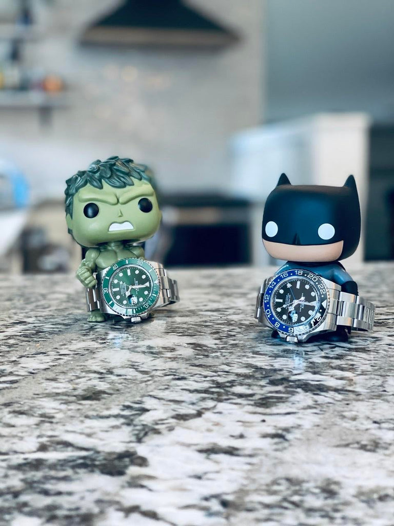 rolex gmt master II batman and green submariner hulk laying on counter top with hulk and batman pop figurines