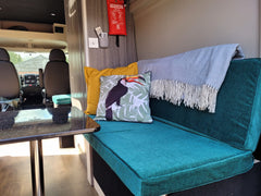 A camper van seat cushion with piping that pulls out to a mattress. The cushion is made up in a hard wearing upholstery fabric in a teal colour. On the seating are are two scatter cushions, one in bright yellow and the other in blues with a toucan on it. You can see through to the front of the van where the two front driving seats are.