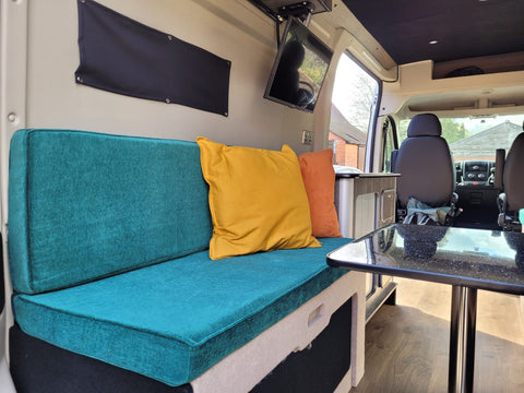 The inside of a recently done camper van, focussed on a bench seat with a back as well which pulls out to a bed for sleeping on. You can see a small table next to the seat as well as a bit of the view out of the front of the van, with the two front driving seats.