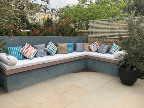 A cosy fenced in London patio garden with a long bench seating area. On top of the bench are three long white outdoor fabric foam cushion pads. At the end of which are round bolster cushions in a matching white outdoor fabric. There are a selection of brightly coloured scatter throw cushions along the length of the benches.