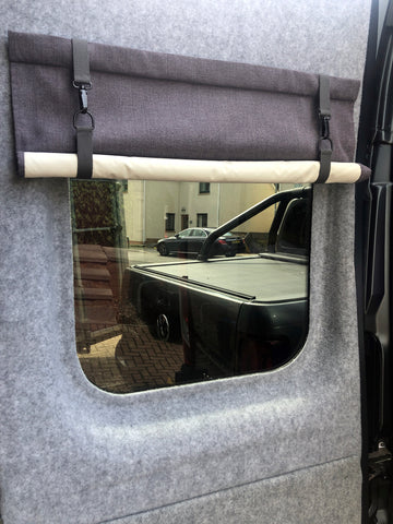 A close up shot of a black out lined blind for a campervan side window. It is held up with a piece of wood and has clasps to keep the blind rolled up. The blind is a medium slate grey with a cream lining.