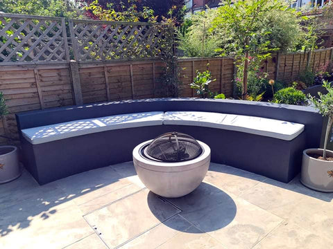 A fire pit on a garden patio. Around the fire pit is a large curved concrete seating area which has two made to measure, accurately shaped foam bench cushions on top, which are amde in water resistant, UV protected fabric in a light grey colour.